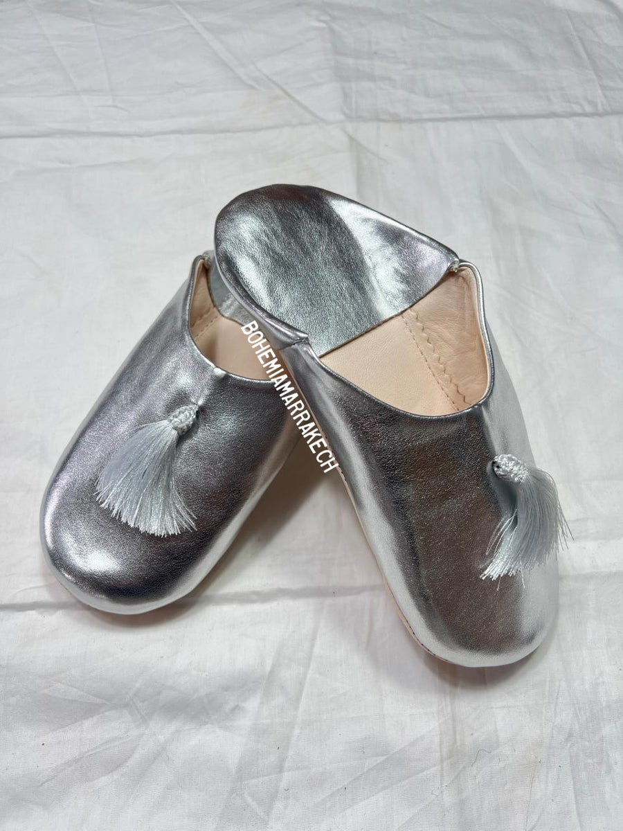 Silver Moroccan babouche slippers with tassel detail on light yellow background