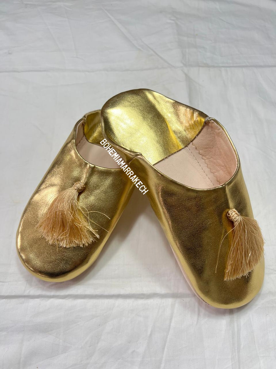 Gold Moroccan babouche slippers with tassel worn on model feet against white background