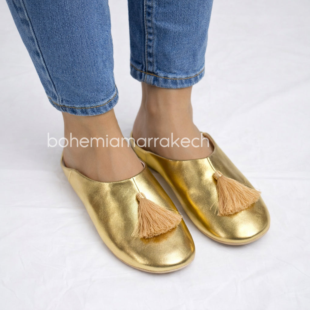 Gold Moroccan babouche slippers with tassel worn on model feet against white background